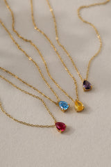 Gold birthstone necklace with two pear-cut stones on a fine cable chain, resting on a pale neutral background