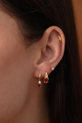 Five gold huggie hoop earrings with pink, clear, green, purple, and blue center gemstones, each flanked by two clear stones, arranged in a row on a soft neutral surface.