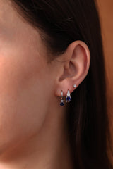 Five silver huggie hoop earrings with red, clear, green, purple, and blue center gemstones, each flanked by two clear stones, arranged in a row on a soft neutral surface.