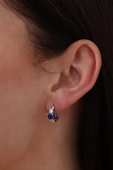 Five silver huggie hoop earrings with red, clear, green, purple, and blue center gemstones, each flanked by two clear stones, arranged in a row on a soft neutral surface.