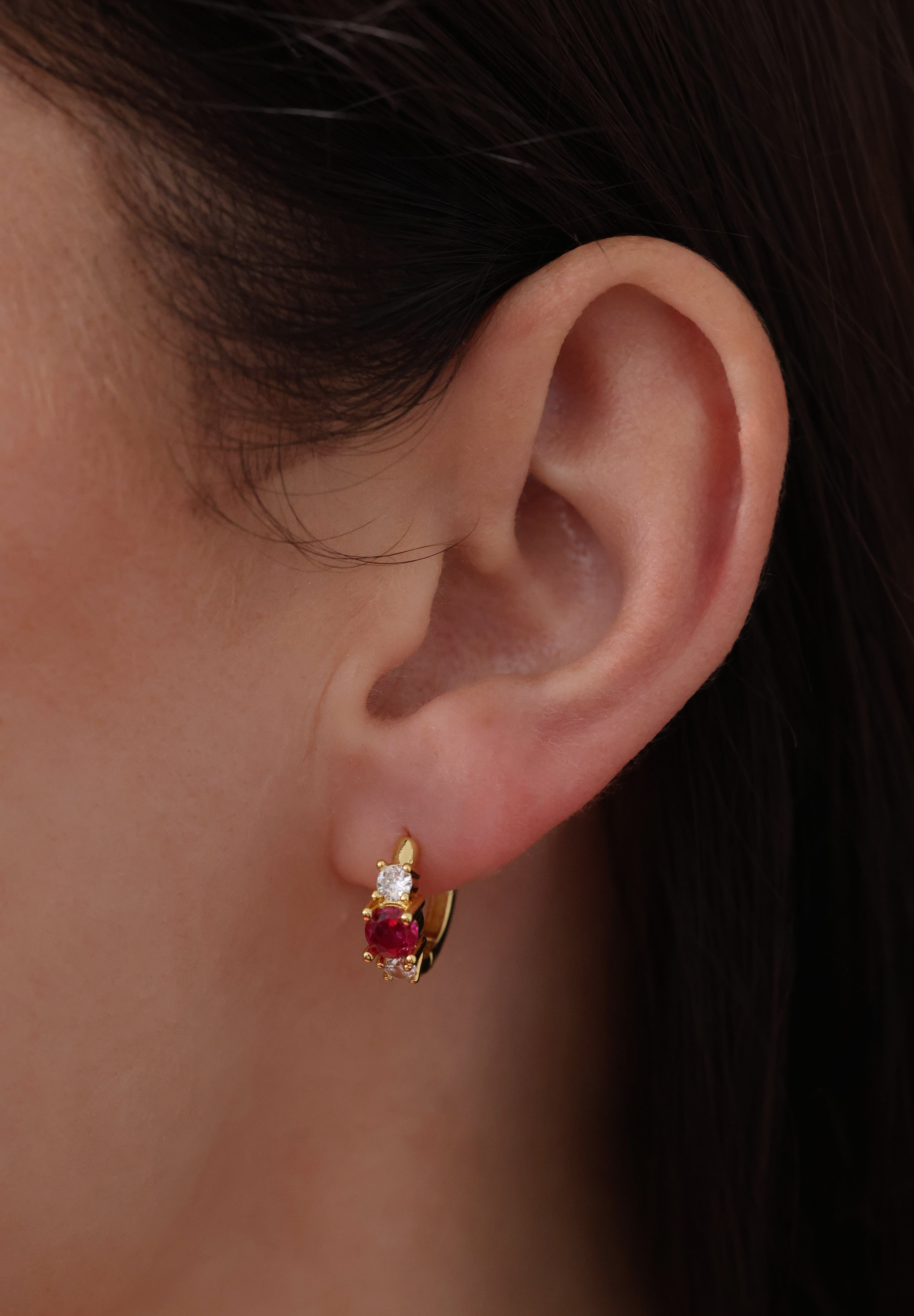 Five gold huggie hoop earrings with pink, clear, green, purple, and blue center gemstones, each flanked by two clear stones, arranged in a row on a soft neutral surface.