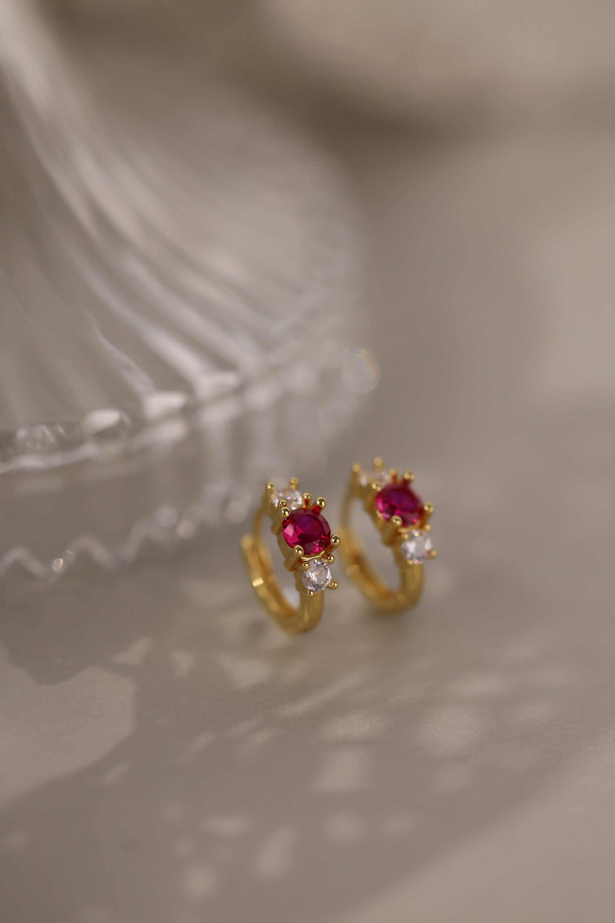 Five gold huggie hoop earrings with pink, clear, green, purple, and blue center gemstones, each flanked by two clear stones, arranged in a row on a soft neutral surface.