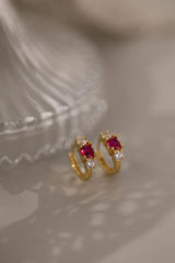 Five gold huggie hoop earrings with pink, clear, green, purple, and blue center gemstones, each flanked by two clear stones, arranged in a row on a soft neutral surface.