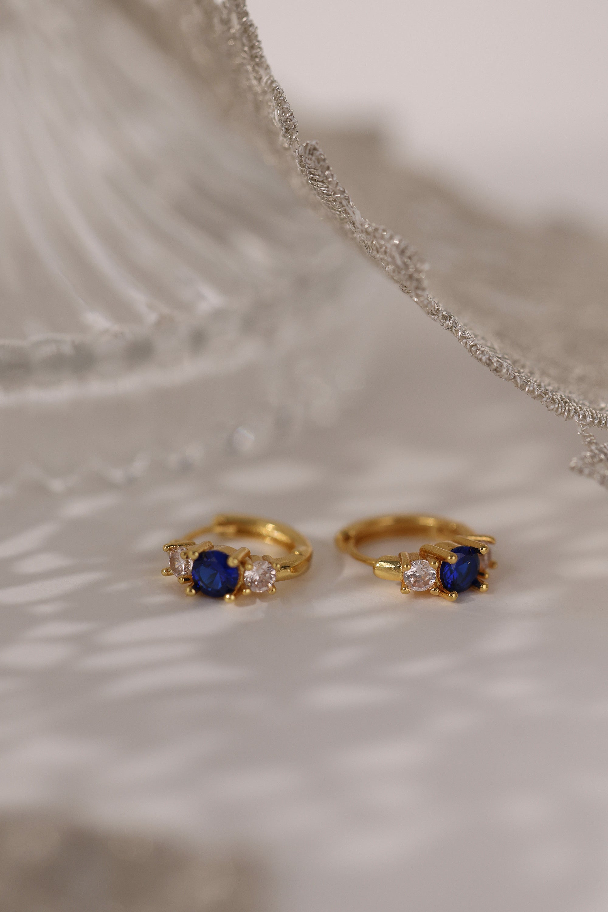 Five gold huggie hoop earrings with pink, clear, green, purple, and blue center gemstones, each flanked by two clear stones, arranged in a row on a soft neutral surface.