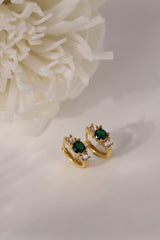 Five gold huggie hoop earrings with red, clear, green, purple, and blue center gemstones, each flanked by two clear stones, arranged in a row on a soft neutral surface.