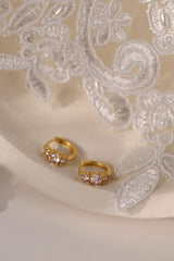 Five gold huggie hoop earrings with pink, clear, green, purple, and blue center gemstones, each flanked by two clear stones, arranged in a row on a soft neutral surface.