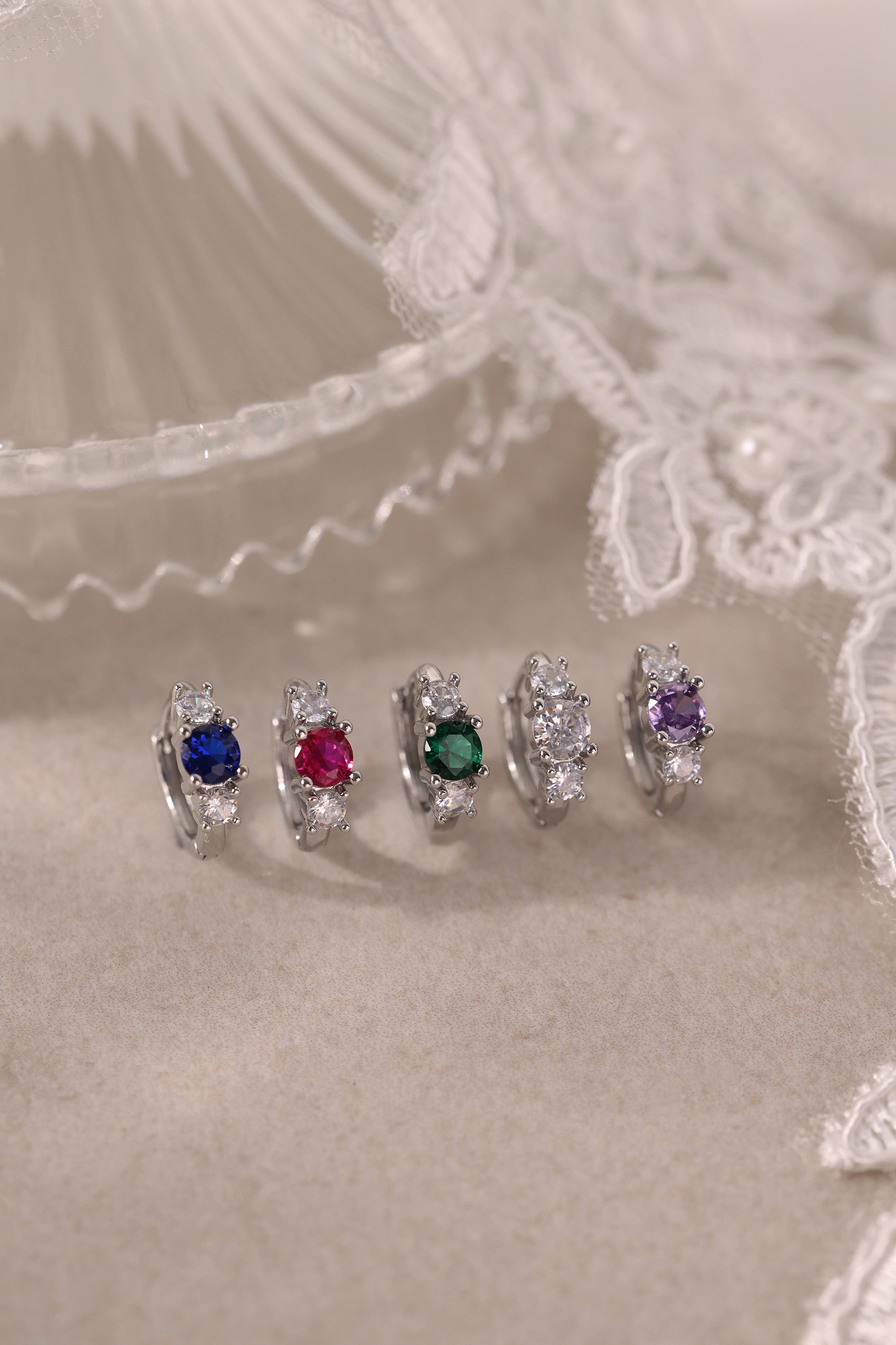Five silver huggie hoop earrings with red, clear, green, purple, and blue center gemstones, each flanked by two clear stones, arranged in a row on a soft neutral surface.