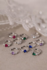 Five silver huggie hoop earrings with red, clear, green, purple, and blue center gemstones, each flanked by two clear stones, arranged in a row on a soft neutral surface.