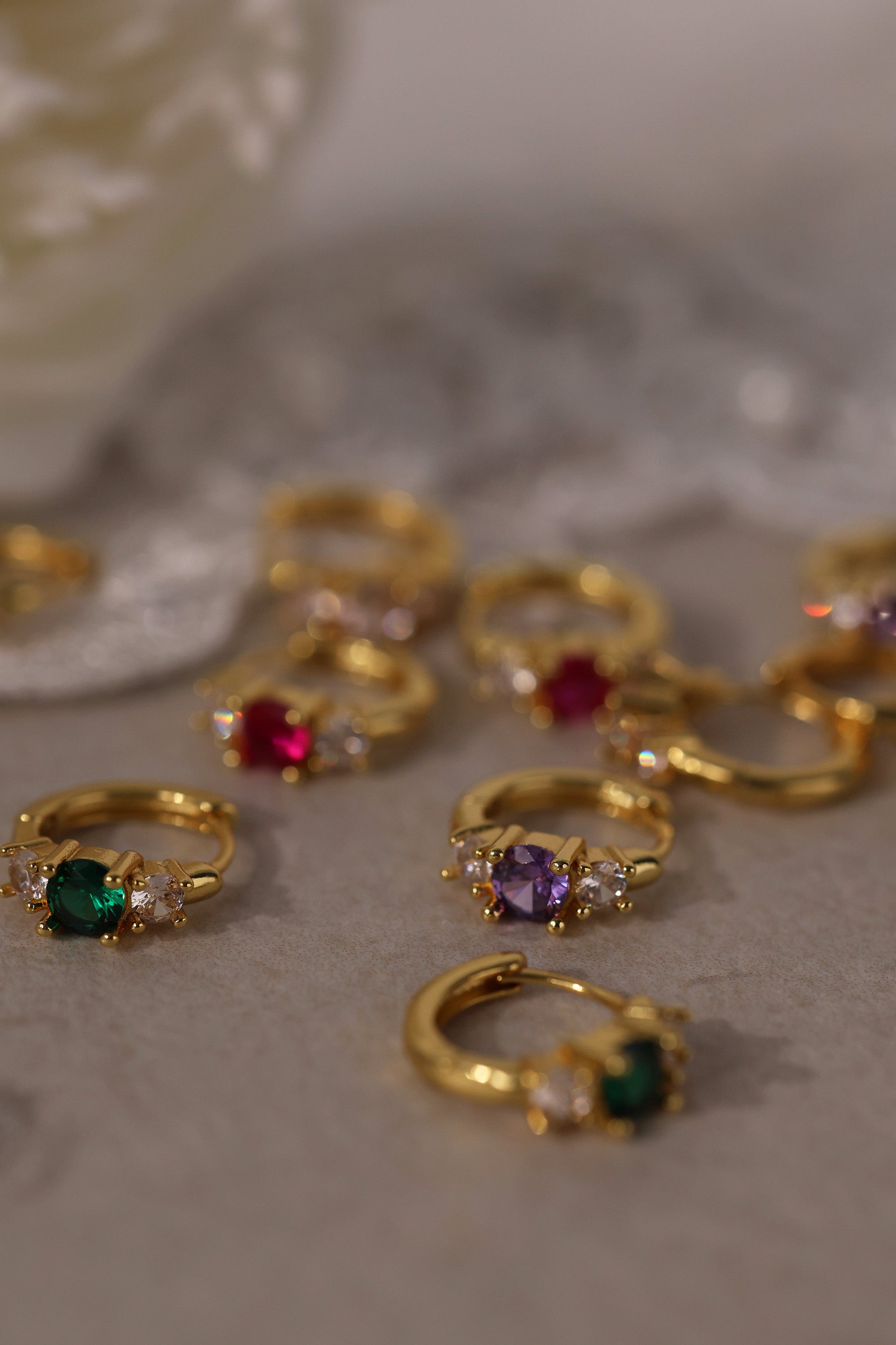 Five gold huggie hoop earrings with pink, clear, green, purple, and blue center gemstones, each flanked by two clear stones, arranged in a row on a soft neutral surface.