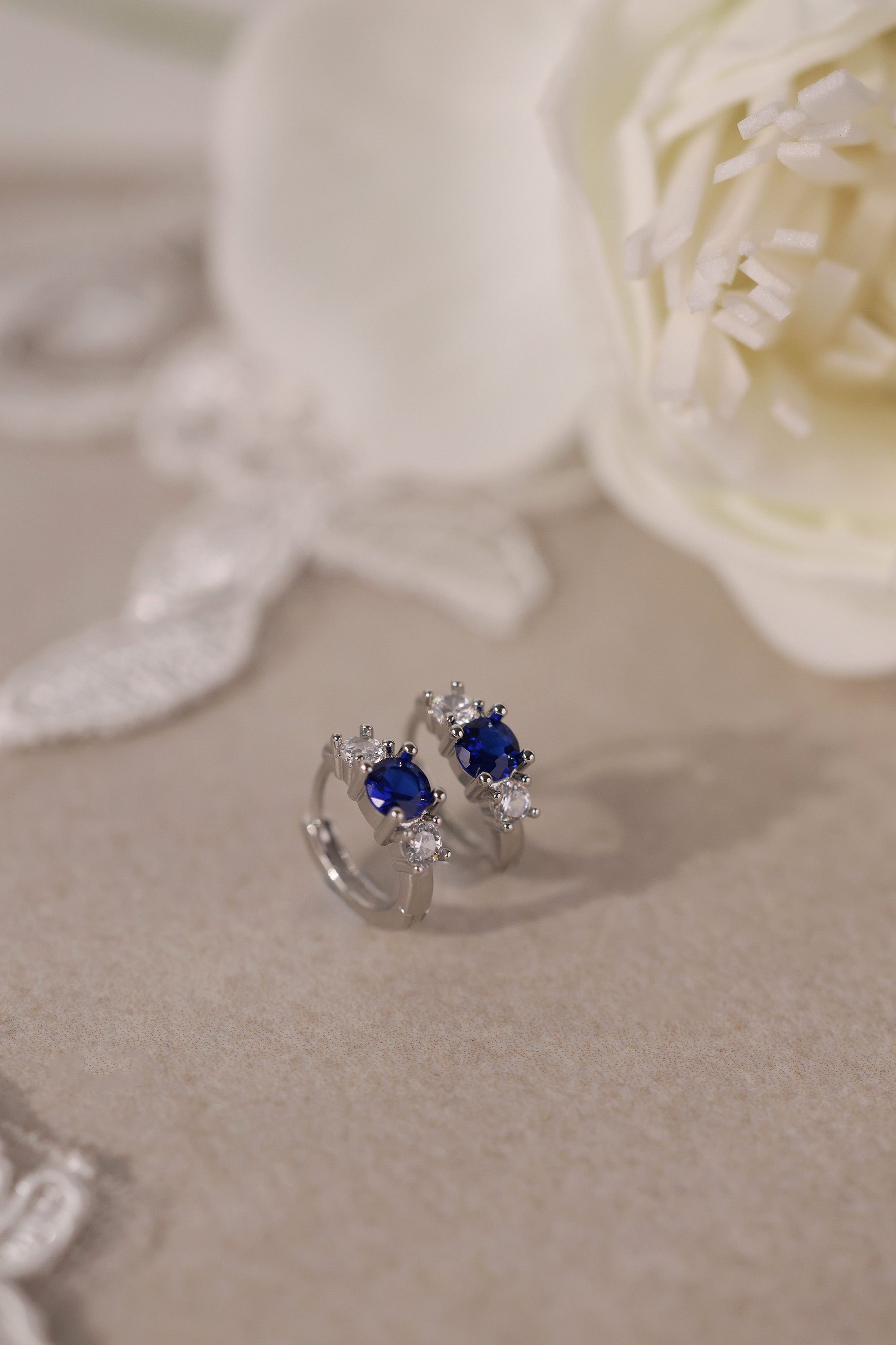 Five silver huggie hoop earrings with red, clear, green, purple, and blue center gemstones, each flanked by two clear stones, arranged in a row on a soft neutral surface.