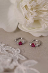 Five silver huggie hoop earrings with red, clear, green, purple, and blue center gemstones, each flanked by two clear stones, arranged in a row on a soft neutral surface.