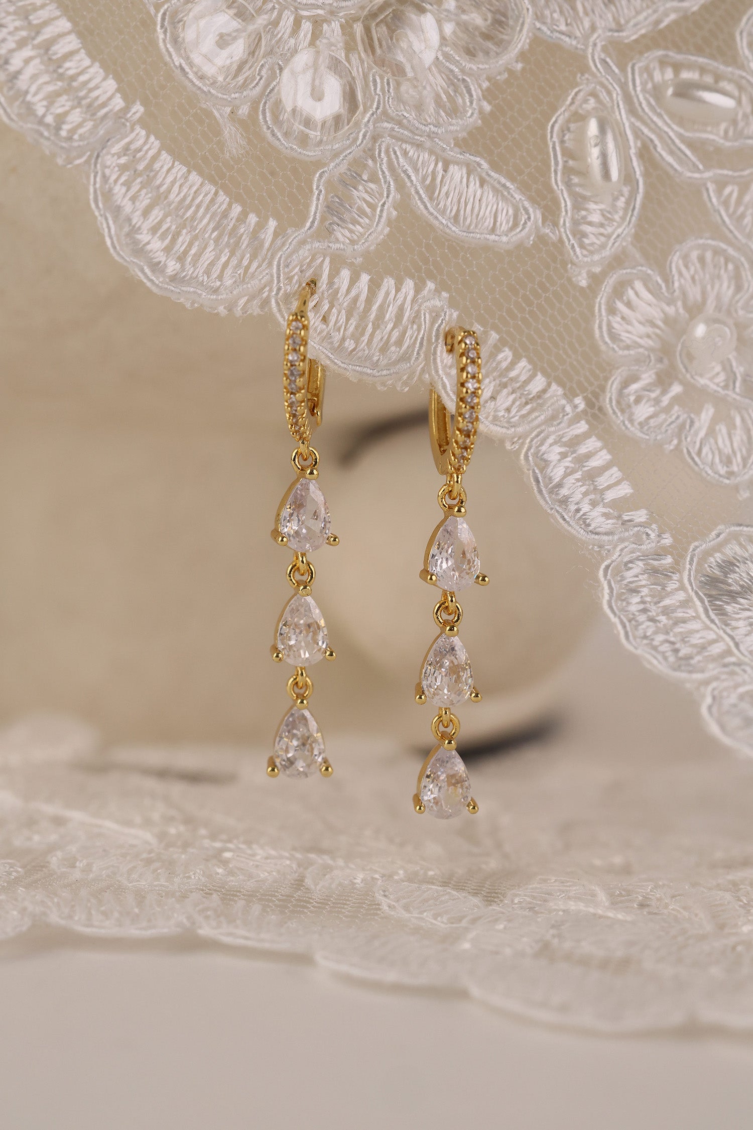 Gold hoop earrings with dangling teardrop gemstones in assorted colors, displayed on embroidered white lace