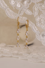 Gold hoop earrings with dangling teardrop gemstones in assorted colors, displayed on embroidered white lace