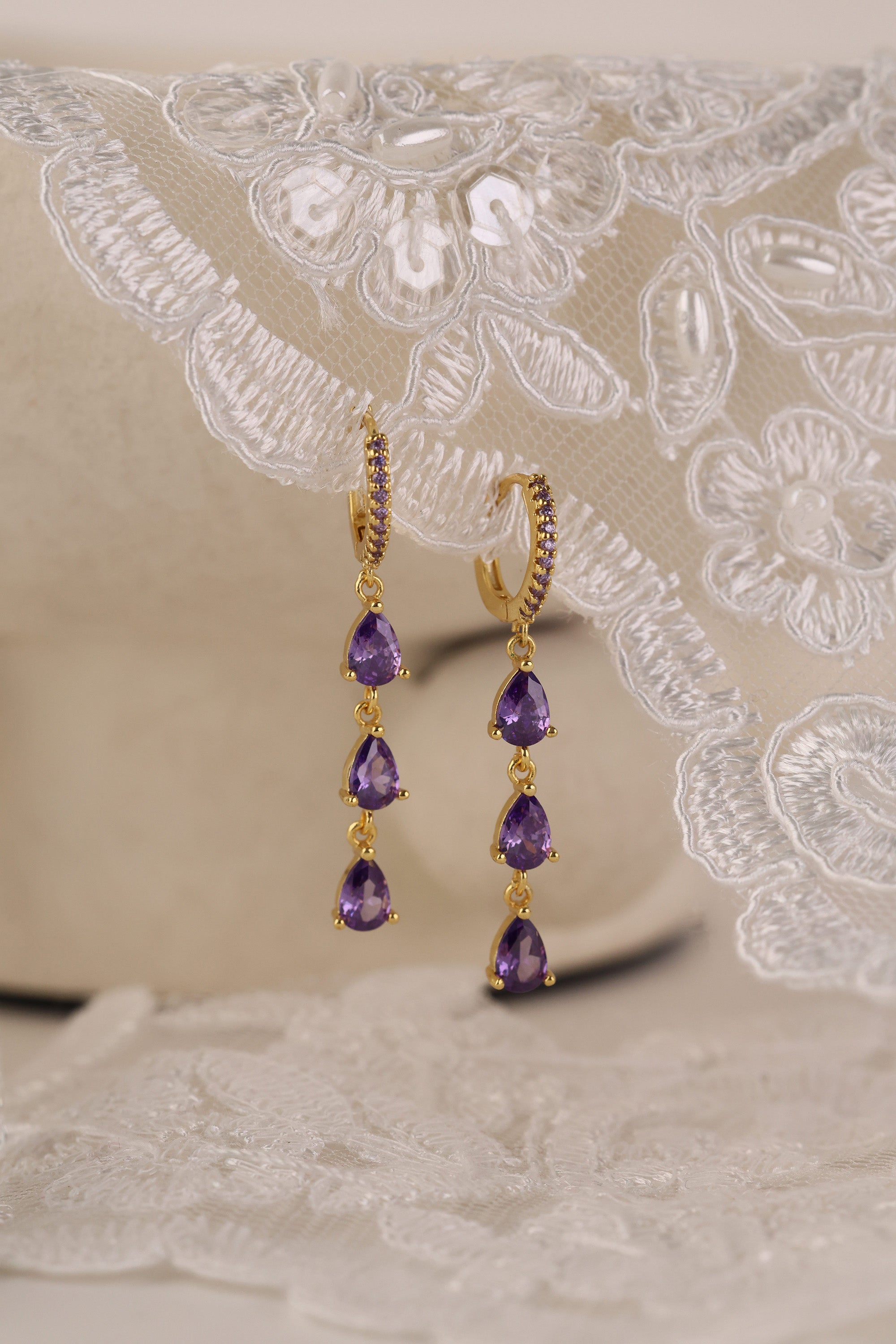 Gold hoop earrings with dangling teardrop gemstones in assorted colors, displayed on embroidered white lace