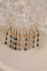 Gold hoop earrings with dangling teardrop gemstones in assorted colors, displayed on embroidered white lace