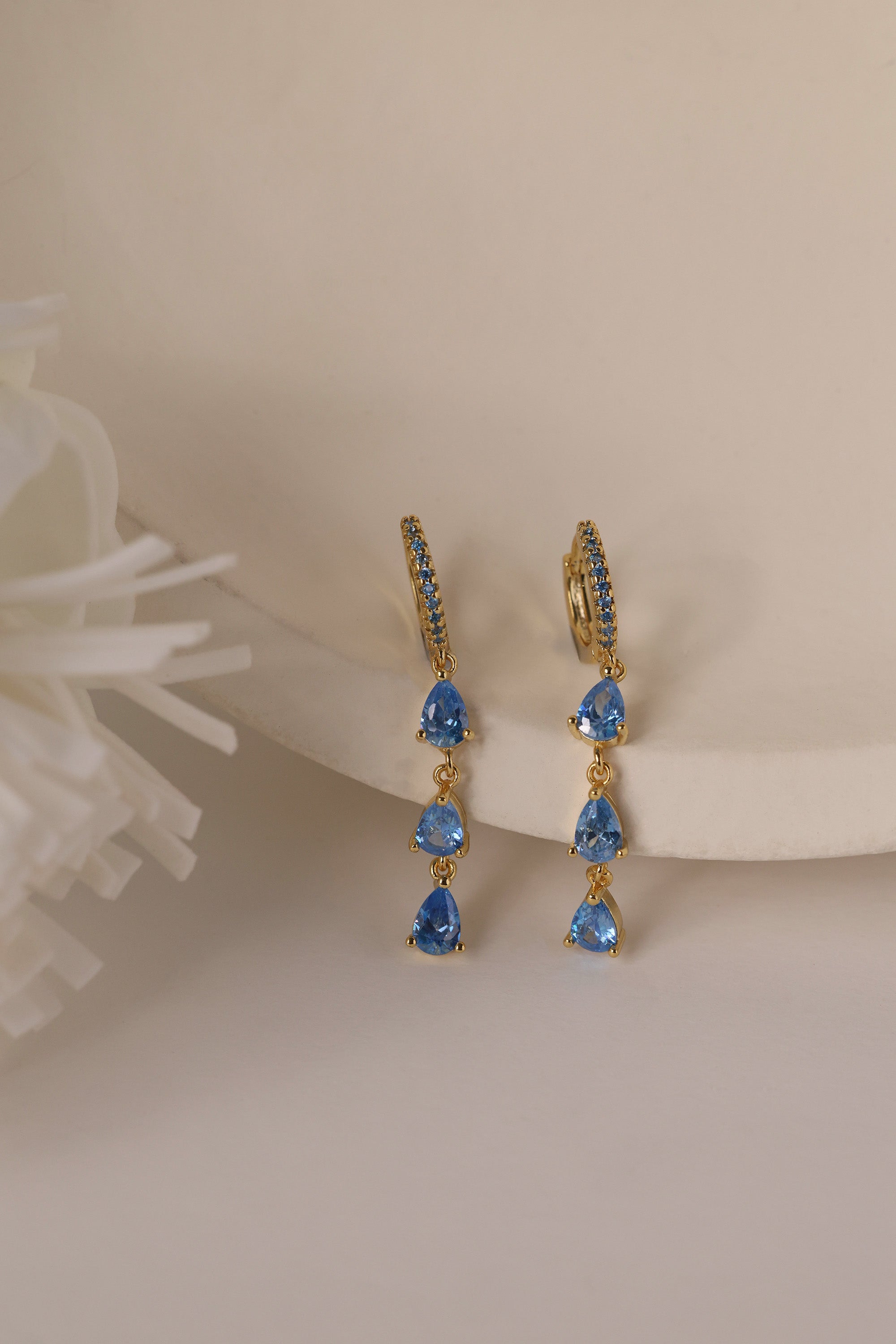 Gold hoop earrings with dangling teardrop gemstones in assorted colors, displayed on embroidered white lace