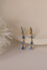 Gold hoop earrings with dangling teardrop gemstones in assorted colors, displayed on embroidered white lace