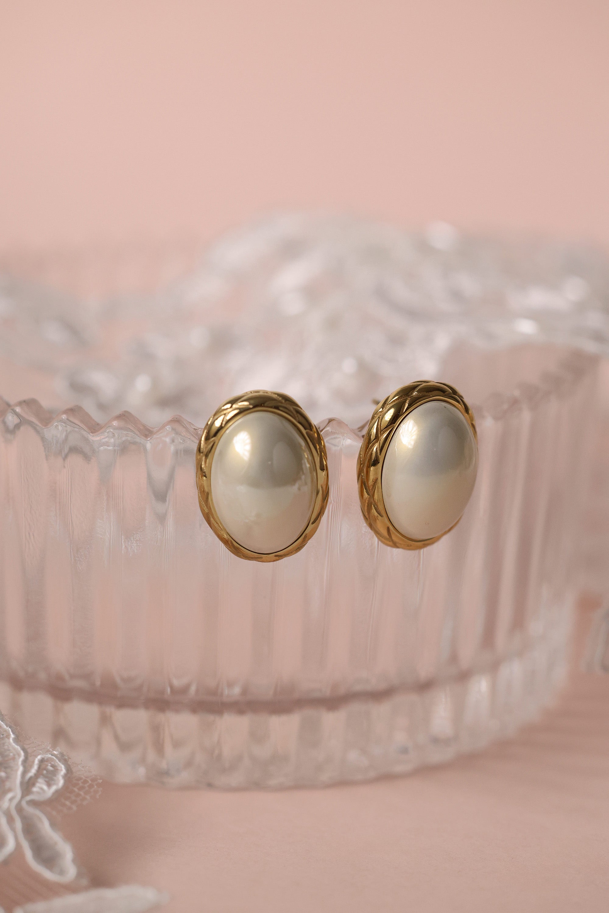 Gold stainless steel oval pearl stud earrings displayed on a glass dish with lace detail.