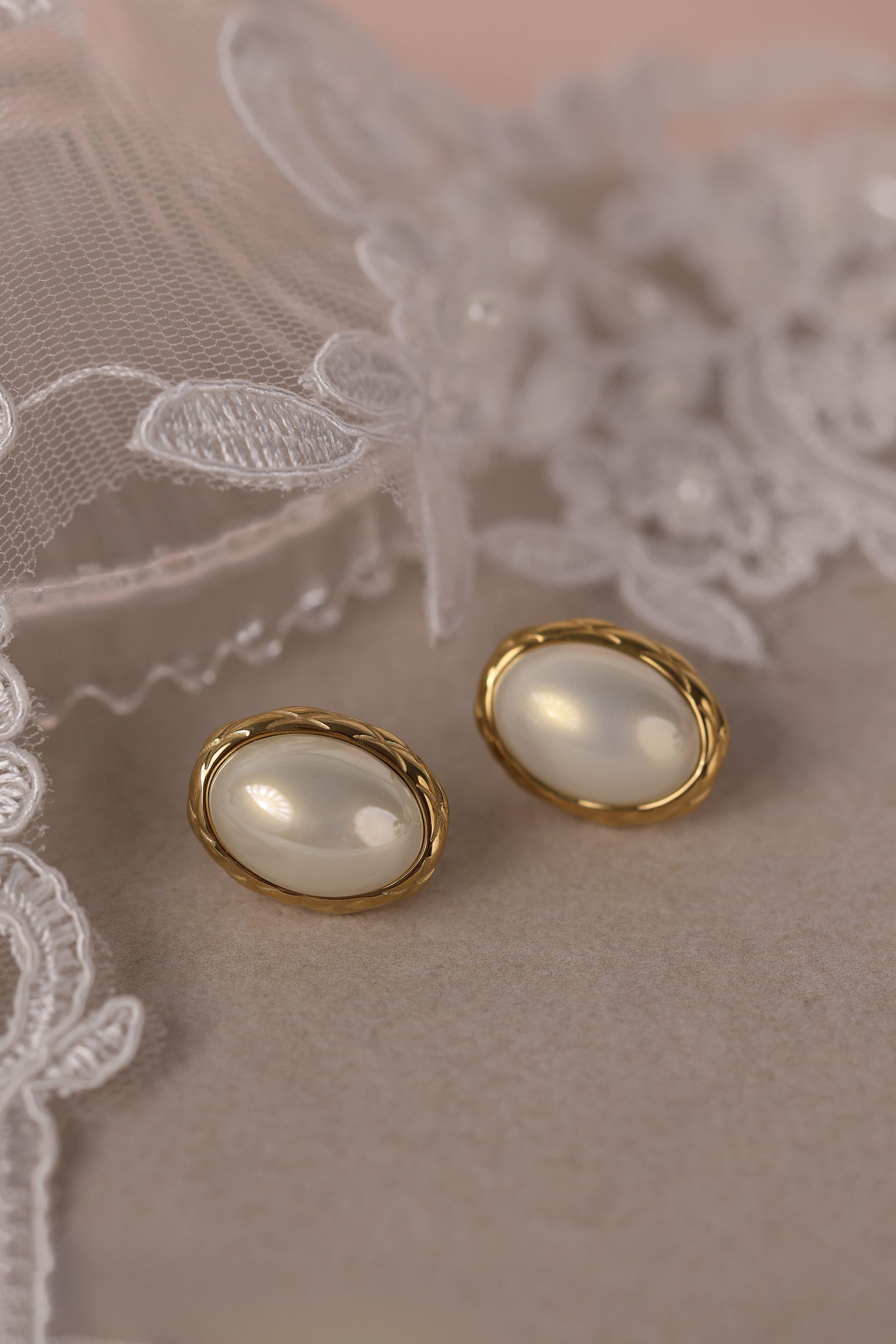 Gold stainless steel oval pearl stud earrings displayed on a glass dish with lace detail.
