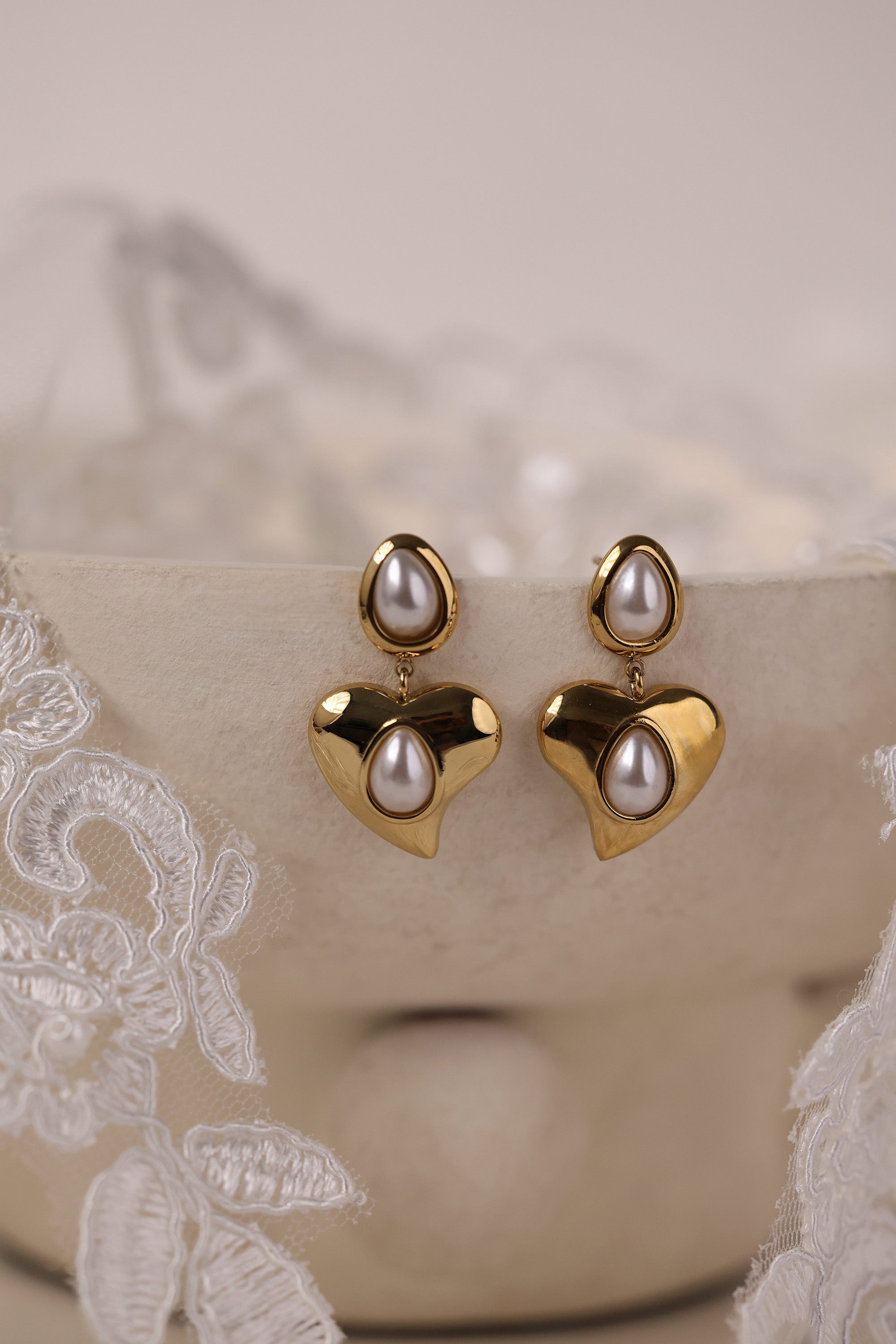 Gold heart stud drop earrings with teardrop pearls displayed on a soft lace background.