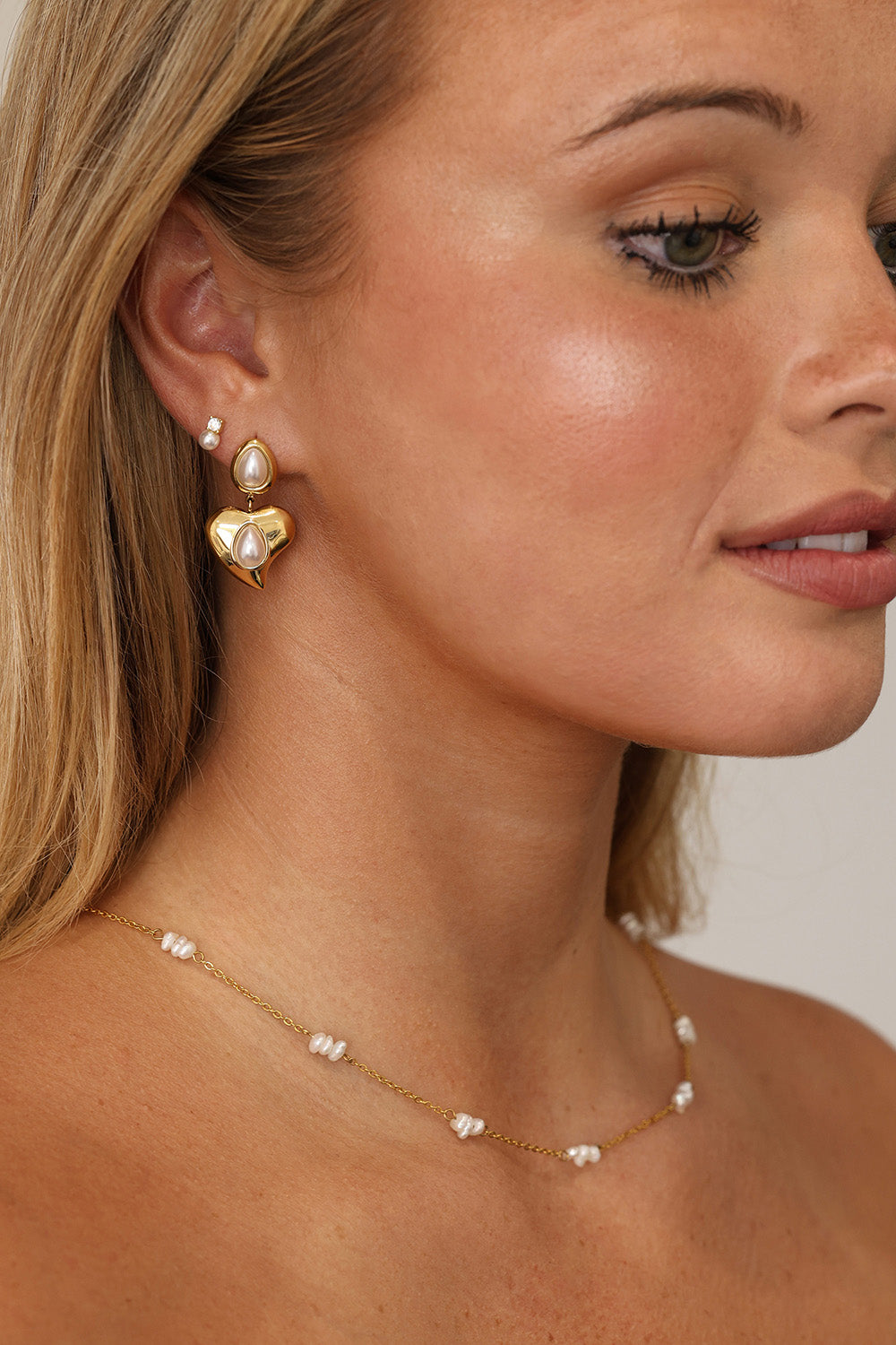 Gold heart stud drop earrings with teardrop pearls displayed on a soft lace background.