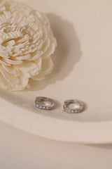 Pair of silver 7mm pavé cubic zirconia huggie hoops on cream beside a dried flower.
