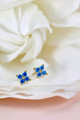 A pair of small gold stud earrings with blue gemstone clusters, resting on the petals of a white rose.