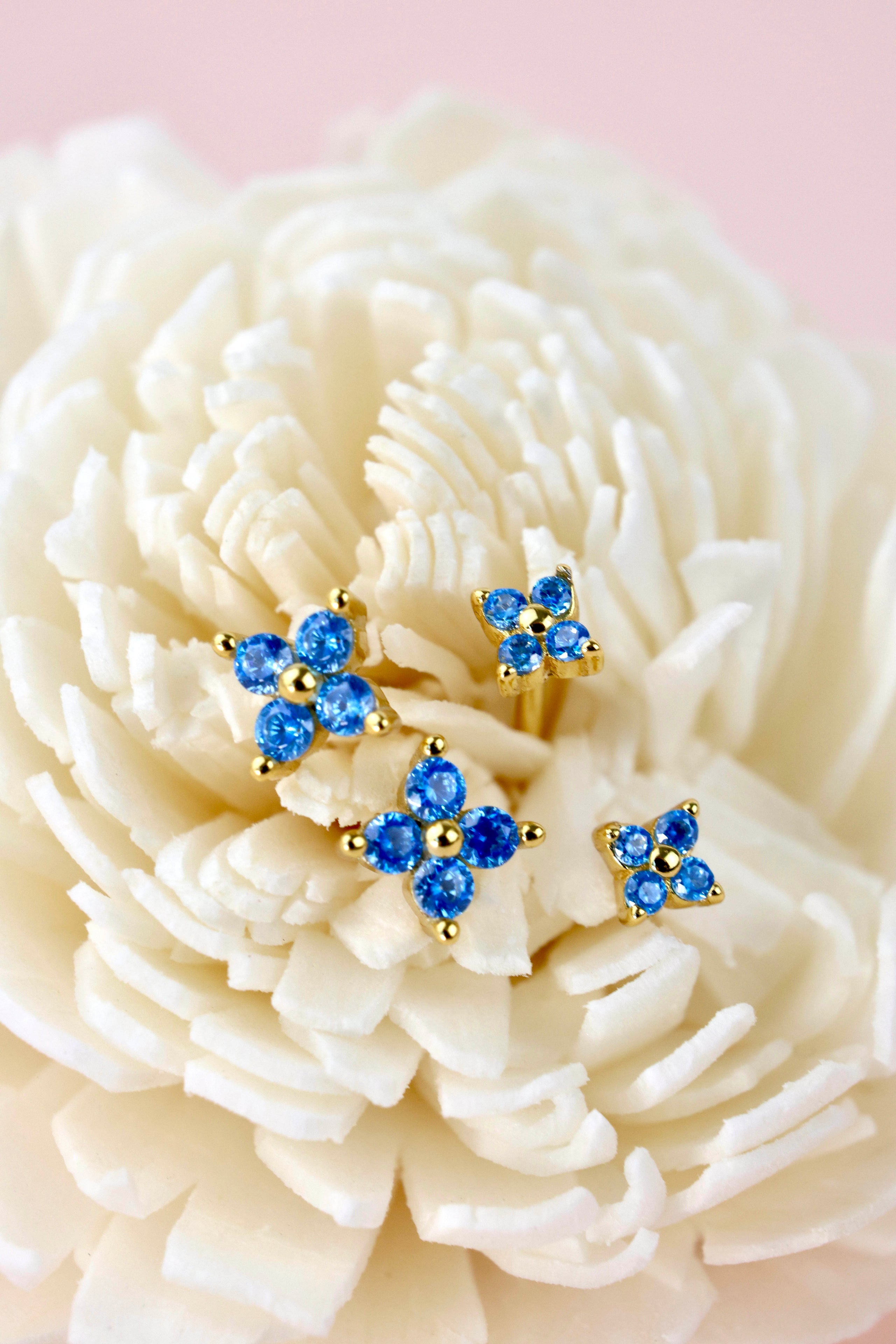 Three pairs of gold-tone stud earrings with blue crystal clusters, displayed on the petals of a large cream-colored flower against a soft pink background.