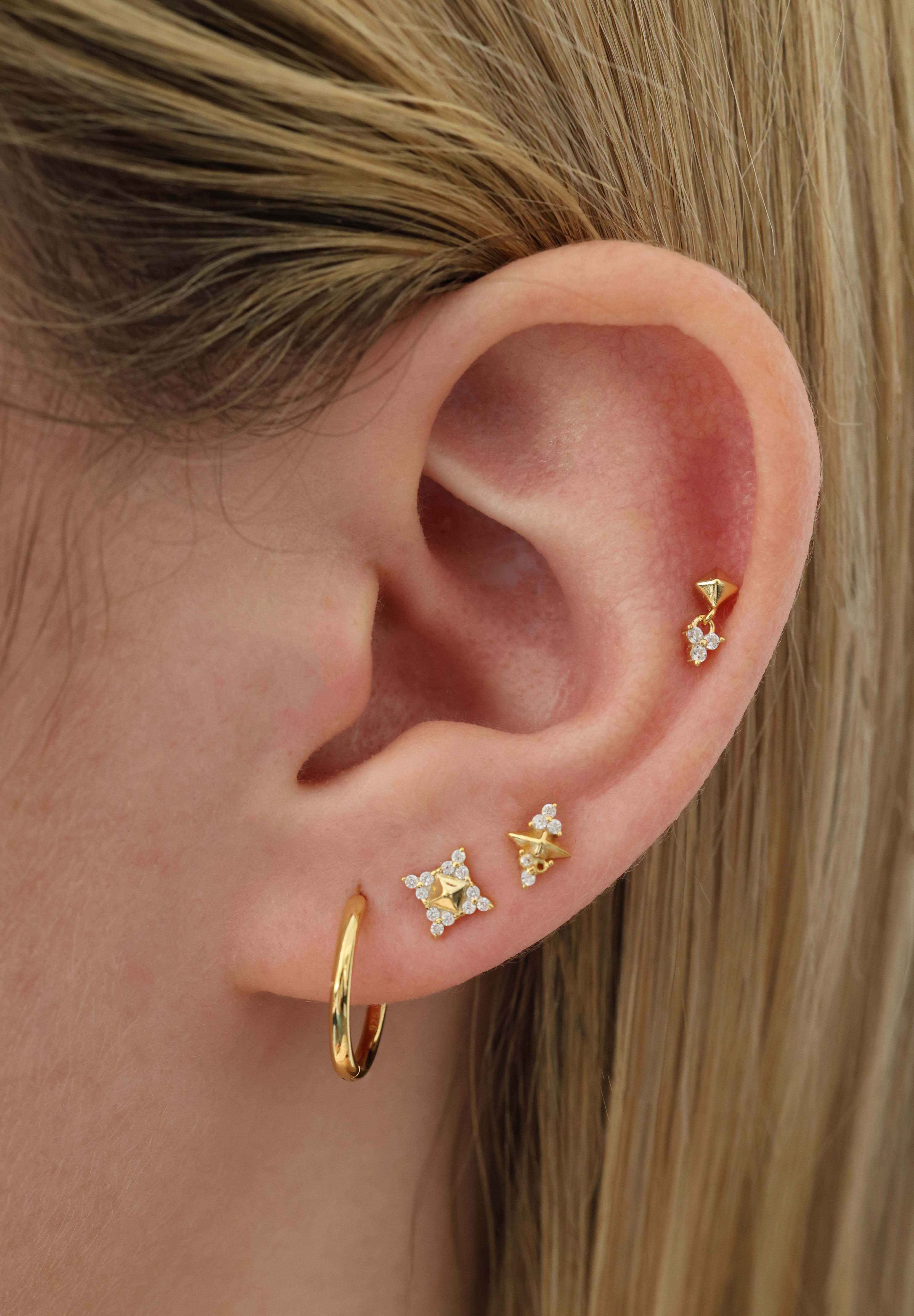 A close-up of a person's ear showcasing multiple gold ear piercings, including a hoop earring, a star-shaped stud, a small geometric stud, and a vertical ear cuff, set against blonde hair.