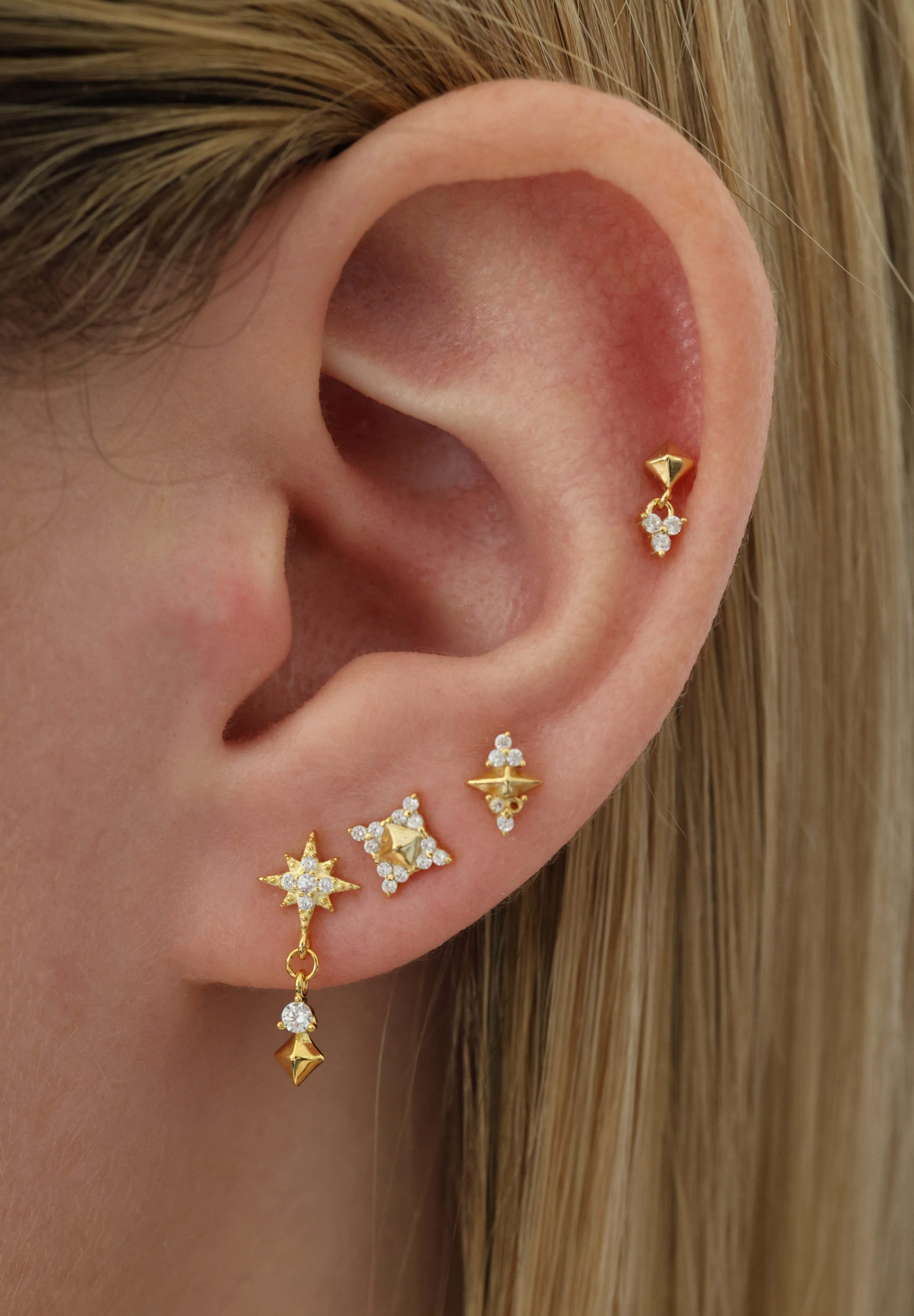 A close-up of a person's ear showcasing a set of gold ear piercings, including a dangle earring with star and lightning motifs, a small star stud, and a geometric stud on the upper lobe.