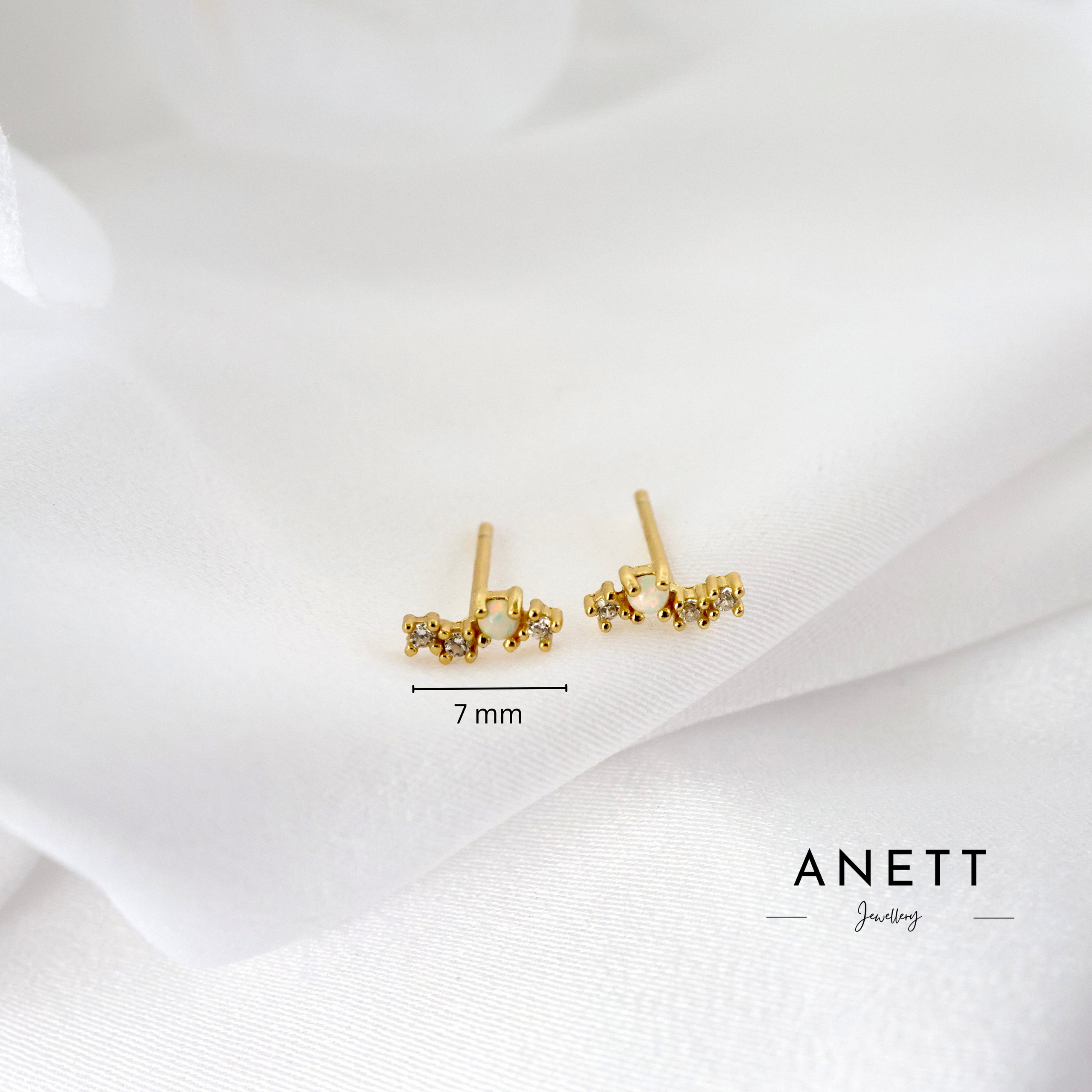A pair of small gold stud earrings with clustered crystal-like details, shown on a soft white fabric background with a 7 mm size marker and the brand name ANETT Jewelry.
