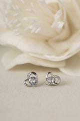 Silver heart stud earrings with a round cubic zirconia centre stone and small CZ accents, shown on a cream background with a white flower