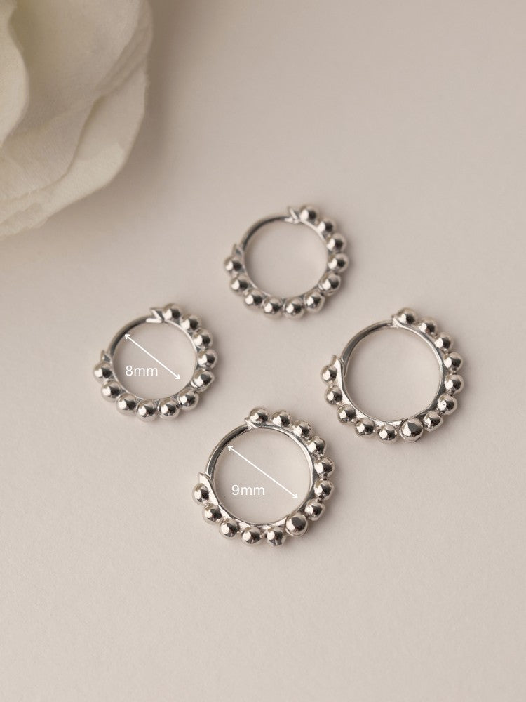 Sterling silver beaded hoop earrings in 8mm and 9mm sizes, featuring polished spherical detailing for a dainty, minimalist look. Displayed on a neutral textured surface with soft floral accents.