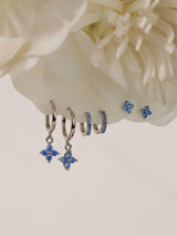 Silver-toned hoop and stud earrings with blue crystal floral motifs, displayed against a soft cream flower backdrop.