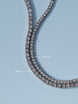 A close-up of a double-row tennis necklace with clear cubic zirconia stones in a silver-toned setting, shown on a light blue background with white text labels indicating 3mm cz and 4mm cz spacing.