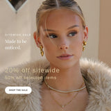 A close-up lifestyle shot of a woman wearing gold jewelry, including a gold dangle earring and layered gold necklaces, styled with a beige fur collar. Promotional text overlays read 'SITEWIDE SALE', 'Made to be noticed.